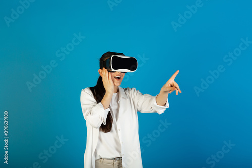 Impressed girl in virtual reality glasses plays a video game and points with her index finger, isolated on a blue background. She's a tech fanatic.