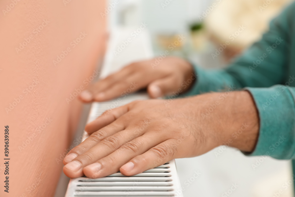 Man warming hands on heating radiator indoors, closeup Man warming hands on heating radiator indoors, closeup