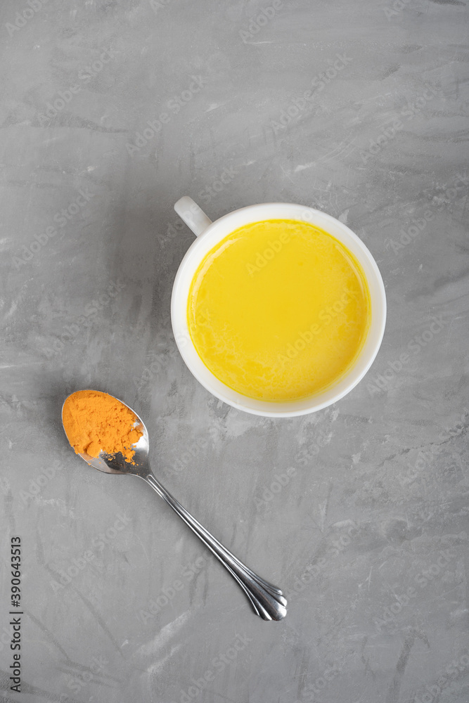 Top view of cup of golden or turmeric milk which is Indian homemade drink used as alternative remedy to boost immunity served with spoon of curcuma powder on gray concrete background. Vertical Top view of cup of golden or turmeric milk which is Indian homemade drink used as alternative remedy to boost immunity served with spoon of curcuma powder on gray concrete background. Vertical