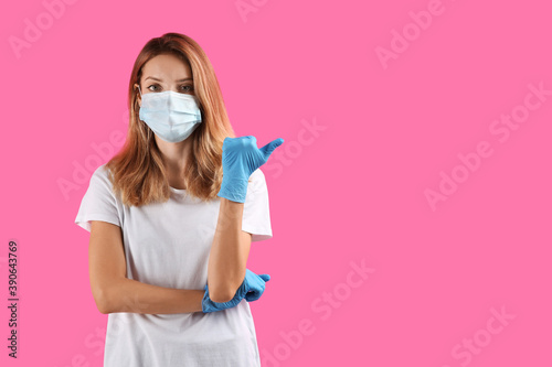 Young woman in medical gloves and protective mask pointing thumb aside on pink background. Space for text