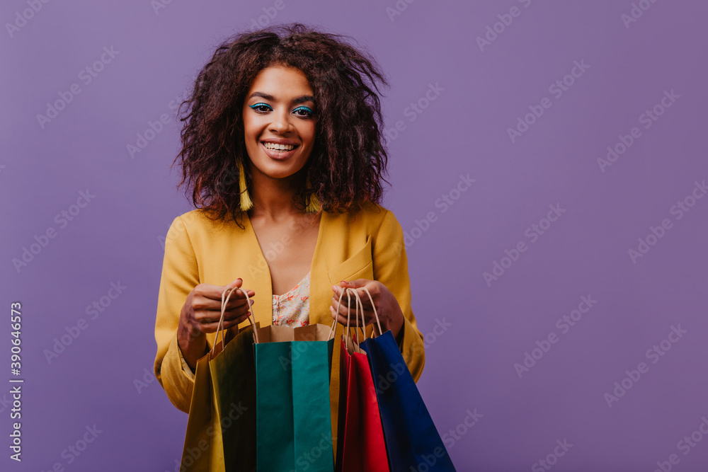 Black cheerful lady posing with colorful paper bags. Charming young woman in yellow jacket having fun after shopping. Black cheerful lady posing with colorful paper bags. Charming young woman in yellow jacket having fun after shopping.