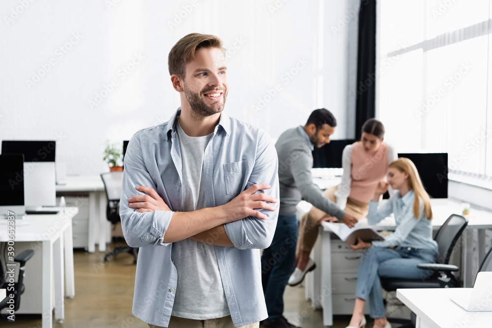 Smiling businessman with crossed arms standing near multicultural colleagues on blurred background in office