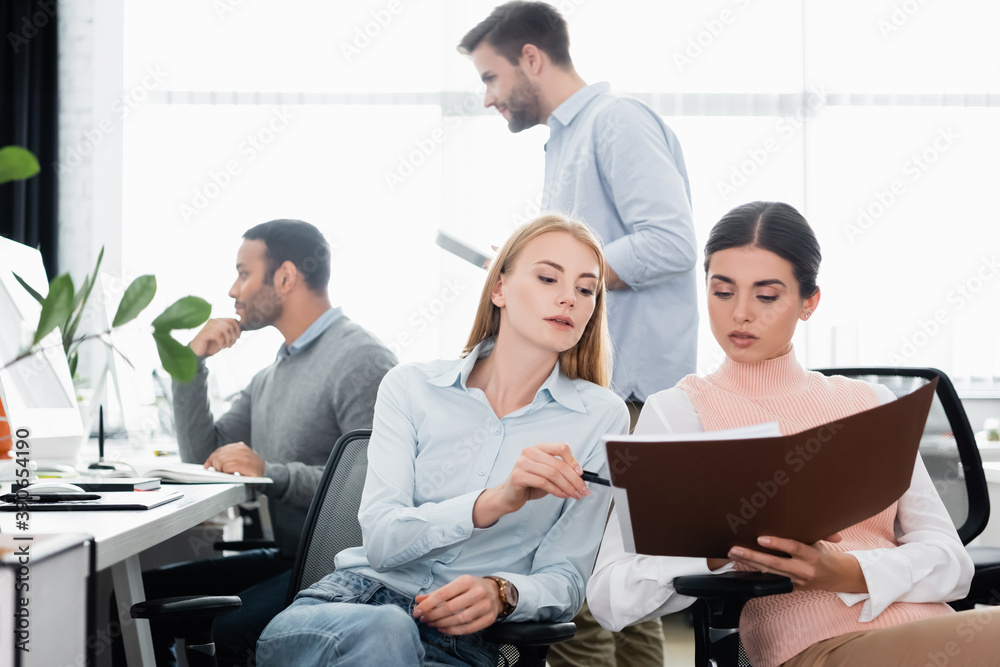 Businesswomen holding pen and paper folder near multicultural colleagues and devices on blurred foreground Businesswomen holding pen and paper folder near multicultural colleagues and devices on blurred foreground