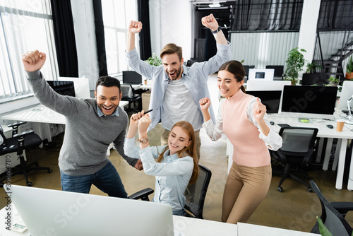 Canvas Print Positive multiethnic businesspeople showing yeah gesture near computer on blurre