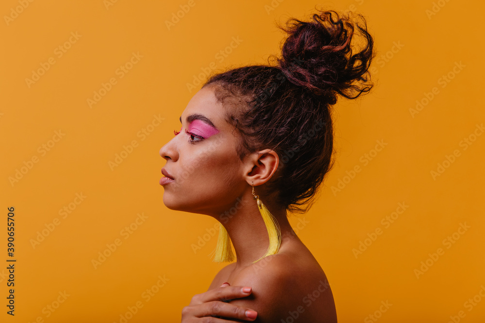Adorable woman with pink makeup spending time in studio. Serious african lady with yellow earrings posing on orange background. Adorable woman with pink makeup spending time in studio. Serious african lady with yellow earrings posing on orange background.