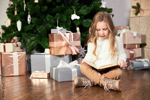 A girl in white eco clothes sits next to a Christmas tree, presents and reads a book