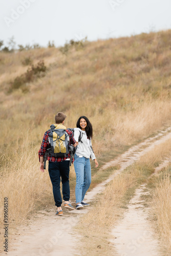 Smiling african american woman looking at boyfriend with backpack while walking on path during trip
