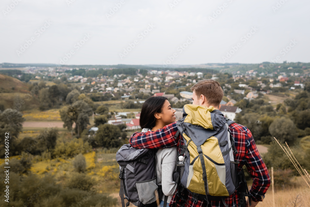 Back view of man embracing african american girlfriend with backpack during trip outdoors