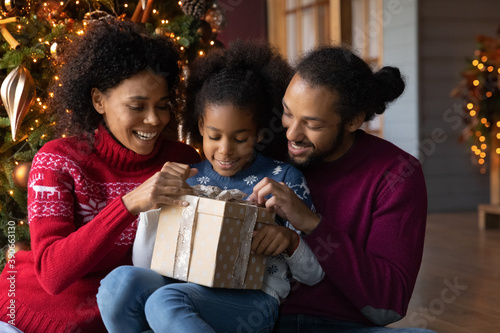 Close up happy African American family unpacking Christmas gift, smiling mother and father with adorable little daughter unwrapping present box, sitting near festive tree at home, winter holiday
