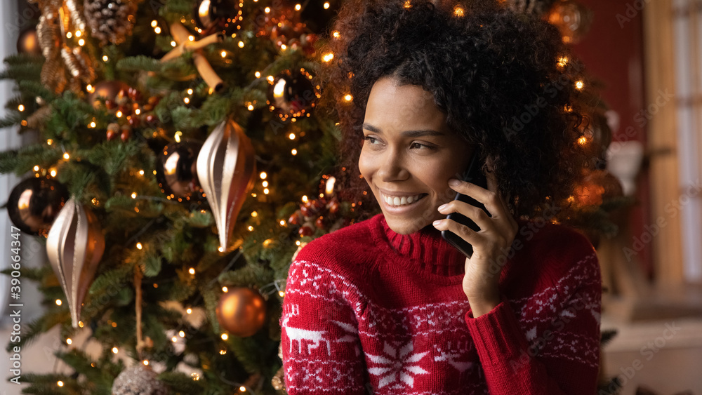Close up smiling African American young woman talking on phone, sitting near festive Christmas tree at home, happy female holding smartphone, chatting with friends or relatives, congratulating - obrazy, fototapety, plakaty 