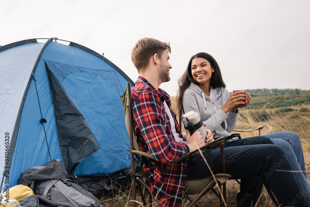 Smiling interracial couple smiling while holding cup and thermos near tent on lawn Smiling interracial couple smiling while holding cup and thermos near tent on lawn