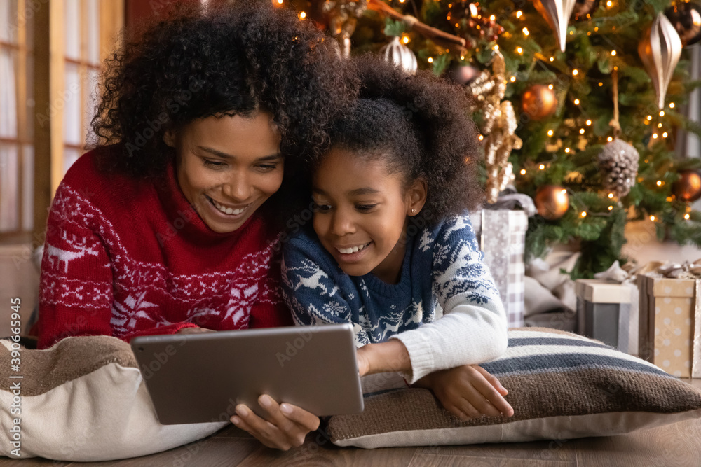 Close up happy African American woman with daughter using tablet together, lying on soft pillows near Christmas tree at home, happy family shopping online, choosing gifts, enjoying winter holiday - obrazy, fototapety, plakaty 