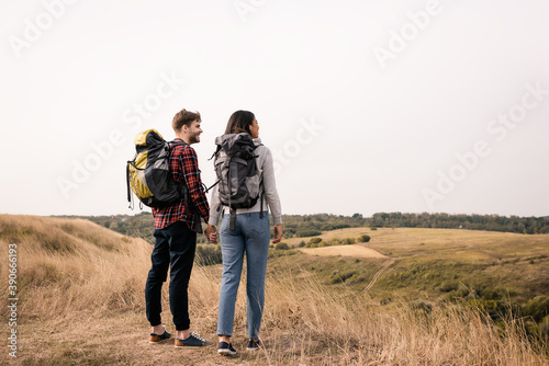 Smiling couple of multiethnic hikers holding hands while looking at landscape on blurred background