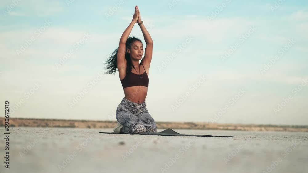 A beautiful young african american woman wearing sportswear is doing yoga exercises sitting on the yoga mat outside at the beach