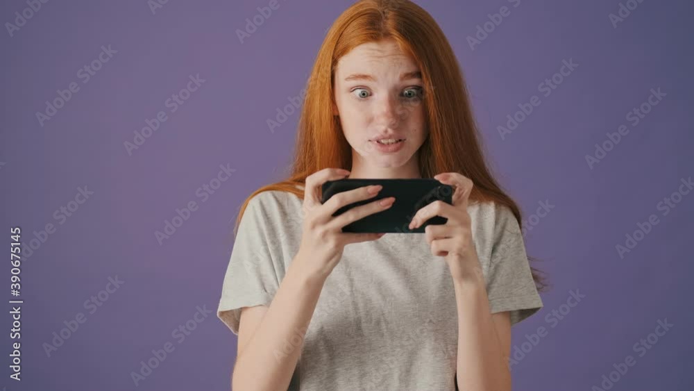 A beautiful young woman is playing games on her smartphone standing isolated over a violet background A beautiful young woman is playing games on her smartphone standing isolated over a violet background
