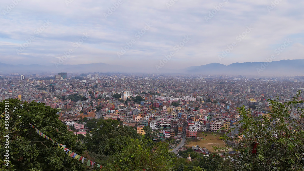 Panorama view of densely populated city center of Kathmandu, capital of Nepal, with visible smog viewed from Buddhist temple complex Swayambhunath with colorful prayer flags, trees and mountains.