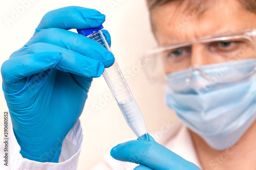 Man wearing mask and safety glasses holds a test tube with blue liquid.Hands close-up, face blurred.Concept of chemical technology, biotechnology, pharmaceuticals.Vaccine against covid-19