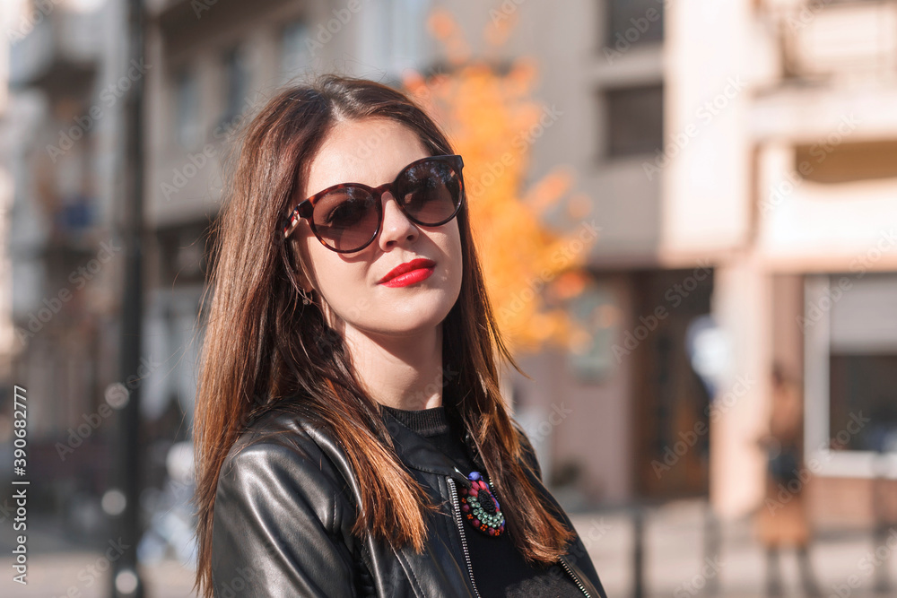 Beautiful woman in a black leather jacket with red lips. Woman in sunglasses looks at the camera. Beautiful woman on a background of autumn city