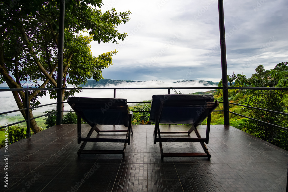 Reclining chairs on the terrace in front of a room with sea fog among trees and mountains in the morning
