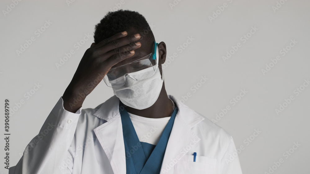 Young African American male doctor in protective eyeglasses and medical mask posing over gray background. Tired after work Young African American male doctor in protective eyeglasses and medical mask posing over gray background. Tired after work