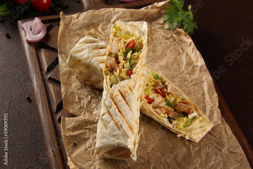 Shawarma with meat and vegetables. Flatbread with chicken, pickles, onions, herbs, mustard and sauce on a wooden board on brown table. Background image, copy space
