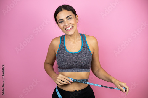 Young beautiful woman wearing sportswear over isolated pink background smiling with a measuring tape around her waist © Irene