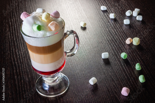 Caffe latte layered with milk in a high drinking glass with marshmallows on top of milk foam. There are marshmallow spread out on the wooden table next to the glass