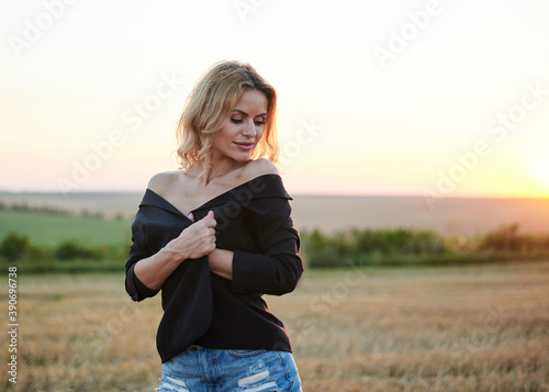 Young blond woman, wearing black jacket and jeans shorts, posing on wheat field on summer evening. Creative stylish three-quarter female portrait during sunset at natural rural landscape.