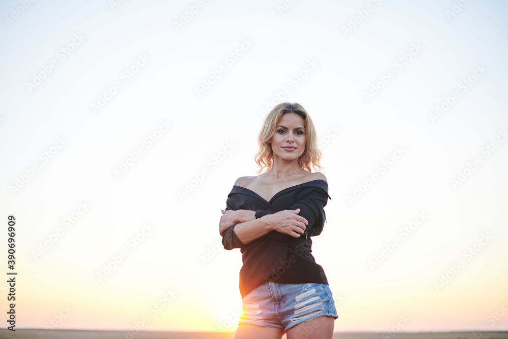 Obraz premium Young blond woman, wearing black jacket and jeans shorts, posing on wheat field on summer evening. Creative stylish three-quarter female portrait during sunset at natural rural landscape.