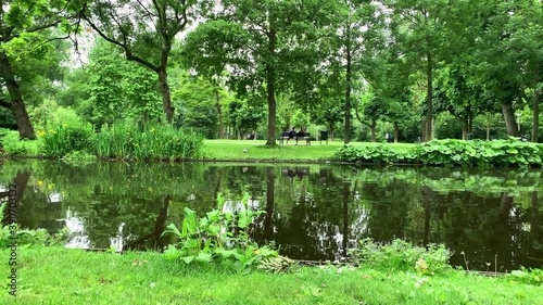Footage of man and woman sitting on a bench, people walking, lake and big trees at Vondelpark in Amsterdam. It is a sunny summer park day.
