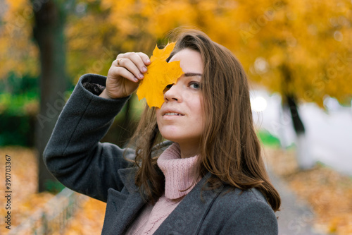 beautiful girl in an autumn coat covers her face with a yellowed maple leaf