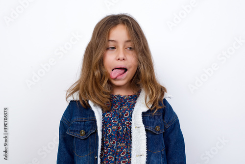 Little caucasian girl with beautiful blue eyes wearing denim jacket standing over isolated white background showing grimace face crossing eyes and showing tongue. Being funny and crazy