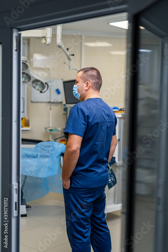 Man doctor wears blue scrubs. Medic standing back to the camera. View from the opened door.