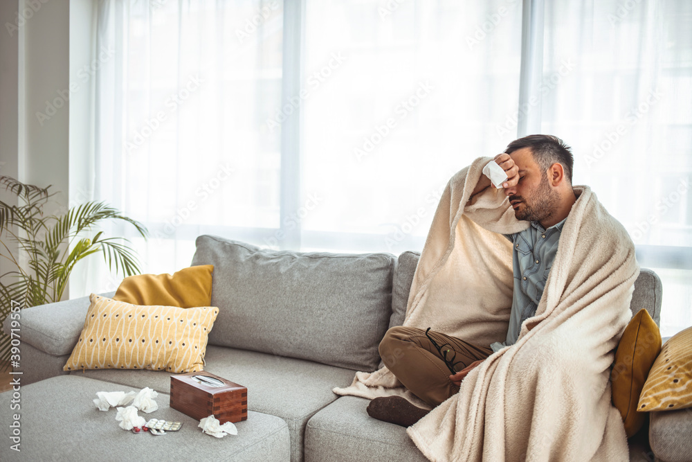 Sickly young man blowing his nose with a tissue in his living room. Young man suffering from cold and coughing. Full length shot of a young man sitting on his bed while feeling unwell at home