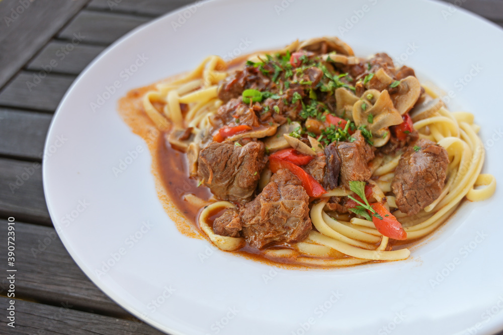 Ragout from beef, red bell peppers and mushrooms with parsley garnish on pasta in a white plate on a dark rustic wooden table, copy space, selected focus