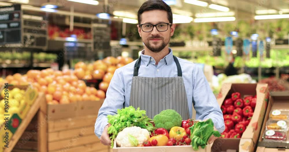 Close up portrait of joyful Caucasian male supermarket worker in good mood holding in hands box with fresh organic vegetables, looking at camera and smiling. Young man in glasses with organic food Close up portrait of joyful Caucasian male supermarket worker in good mood holding in hands box with fresh organic vegetables, looking at camera and smiling. Young man in glasses with organic food