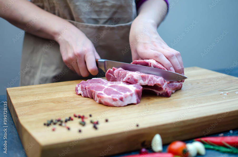 The hostess in the kitchen cuts pork with a knife, prepares food. On a cutting board, spices, chili peppers, rosemary, garlic, cherry tomatoes.