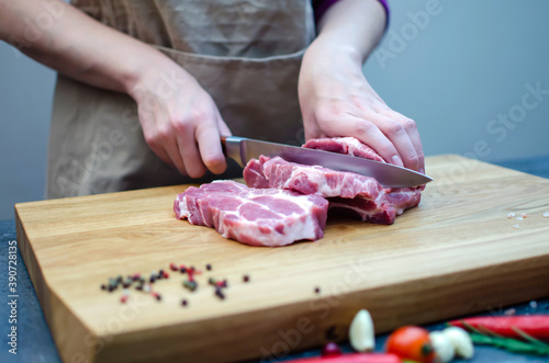 The hostess in the kitchen cuts pork with a knife, prepares food. On a cutting board, spices, chili peppers, rosemary, garlic, cherry tomatoes.