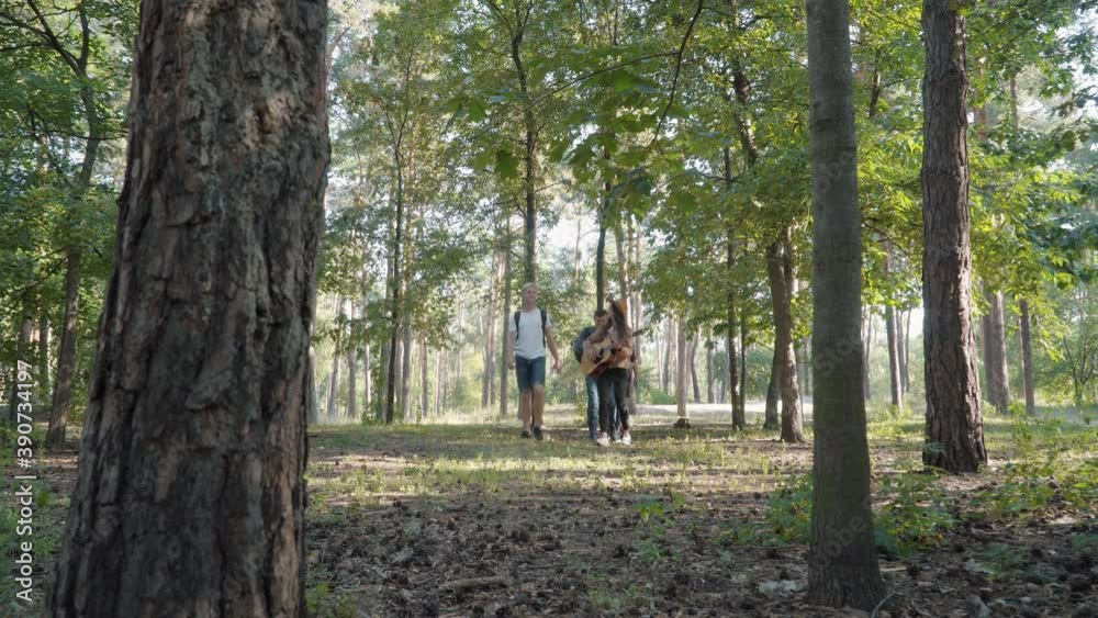 Extreme wide shot of beautiful woman playing guitar as strolling with friends in summer forest. Positive Caucasian group of tourists leaving, handsome man staying to admire nature through binoculars. Extreme wide shot of beautiful woman playing guitar as strolling with friends in summer forest. Positive Caucasian group of tourists leaving, handsome man staying to admire nature through binoculars.