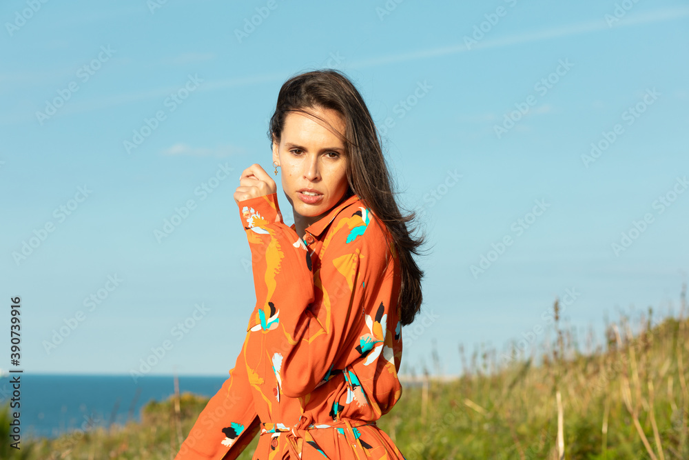 
Young woman with long hair and flowery fashion orange dress posing by tall grass and blue sky. Cute modern European model girl enjoys a walk on the beach. Spring summer fashion