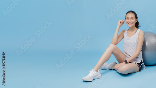 Asian beautiful happy woman sitting on the floor with fit ball after exercise isolated on blue colour background with copy space.Concept of slim and healthy girl workout.