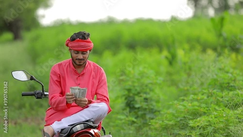 Happy Young indian farmer sitting two wheeler and counting money at field
