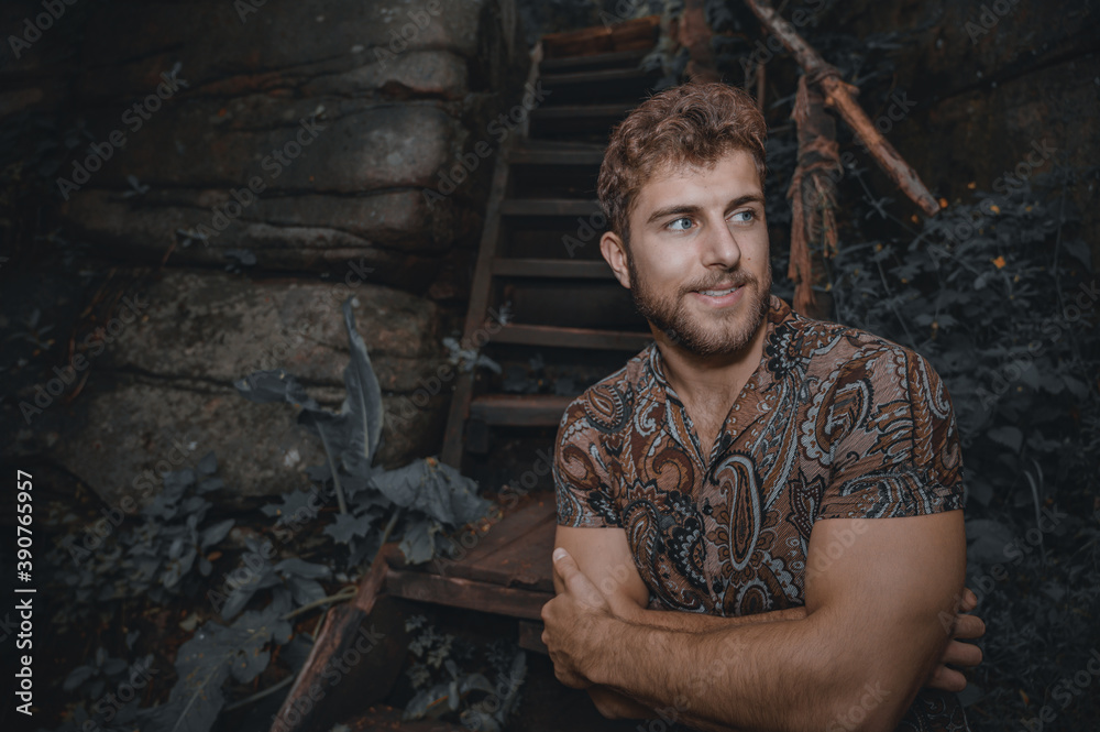 Portrait of strong muscle fit young curly man with brown hazel hair and beard wearing casual motley shirt. In the forest near wooden stairs. Dark natural skin tone, moody. Cheerful male adult journey. Portrait of strong muscle fit young curly man with brown hazel hair and beard wearing casual motley shirt. In the forest near wooden stairs. Dark natural skin tone, moody. Cheerful male adult journey.