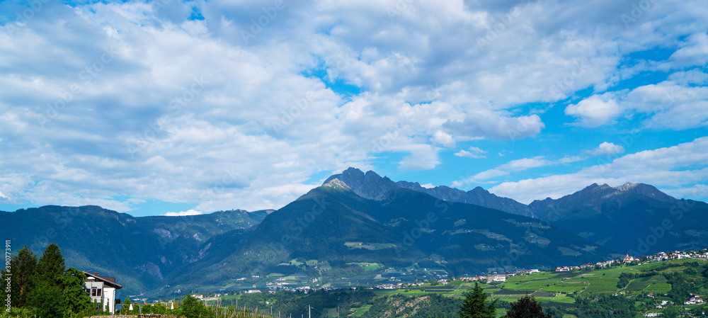 Beautiful alpine landscape with green forest hills, houses and garden. Clouds in blue sky. Agricultural landscape. Picturesque panoramic view of nature.