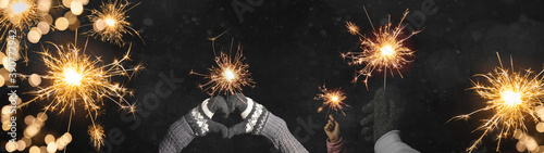 Happy New Year / Silvester / Party background banner panorama greeting card - Young family holding sparkling sparklers in her hands at dark night, with golden bokeh lights and firework