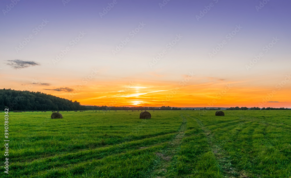 Scenic view at beautiful sunset in green shiny field with hay stacks, bright cloudy sky, country road and golden sun rays with glow, summer valley landscape