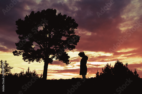 silhouette of a woman standing by a tree outdoors against a sunset background