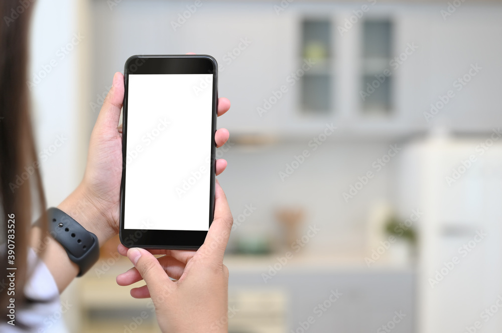 Mockup image of hands showing black mobile phone with blank white screen on blur kitchen background.