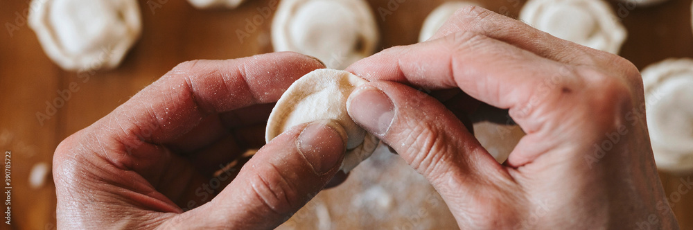 hands of senior man cooking and molding small homemade uncooked dumplings with meat on kitchen table. national traditional Russian cuisine. do it yourself. top view. banner