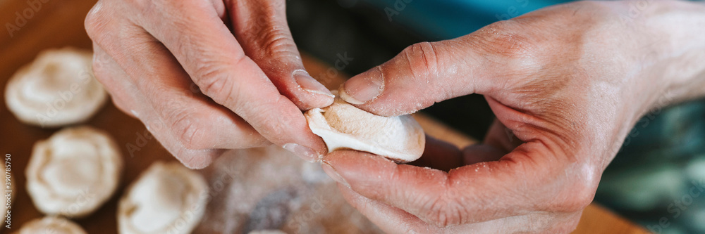 Fototapeta premium hands of senior man cooking and molding small homemade uncooked dumplings with meat on kitchen table. national traditional Russian cuisine. do it yourself. top view. banner